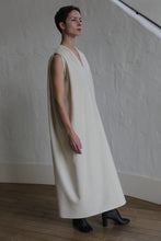 Load image into Gallery viewer, A person with short hair wears The Winter Dress | Cashmere Undyed, a sleeveless, loose-fitting cream dress, paired with black ankle boots, standing indoors on a wooden floor against a textured light wall.