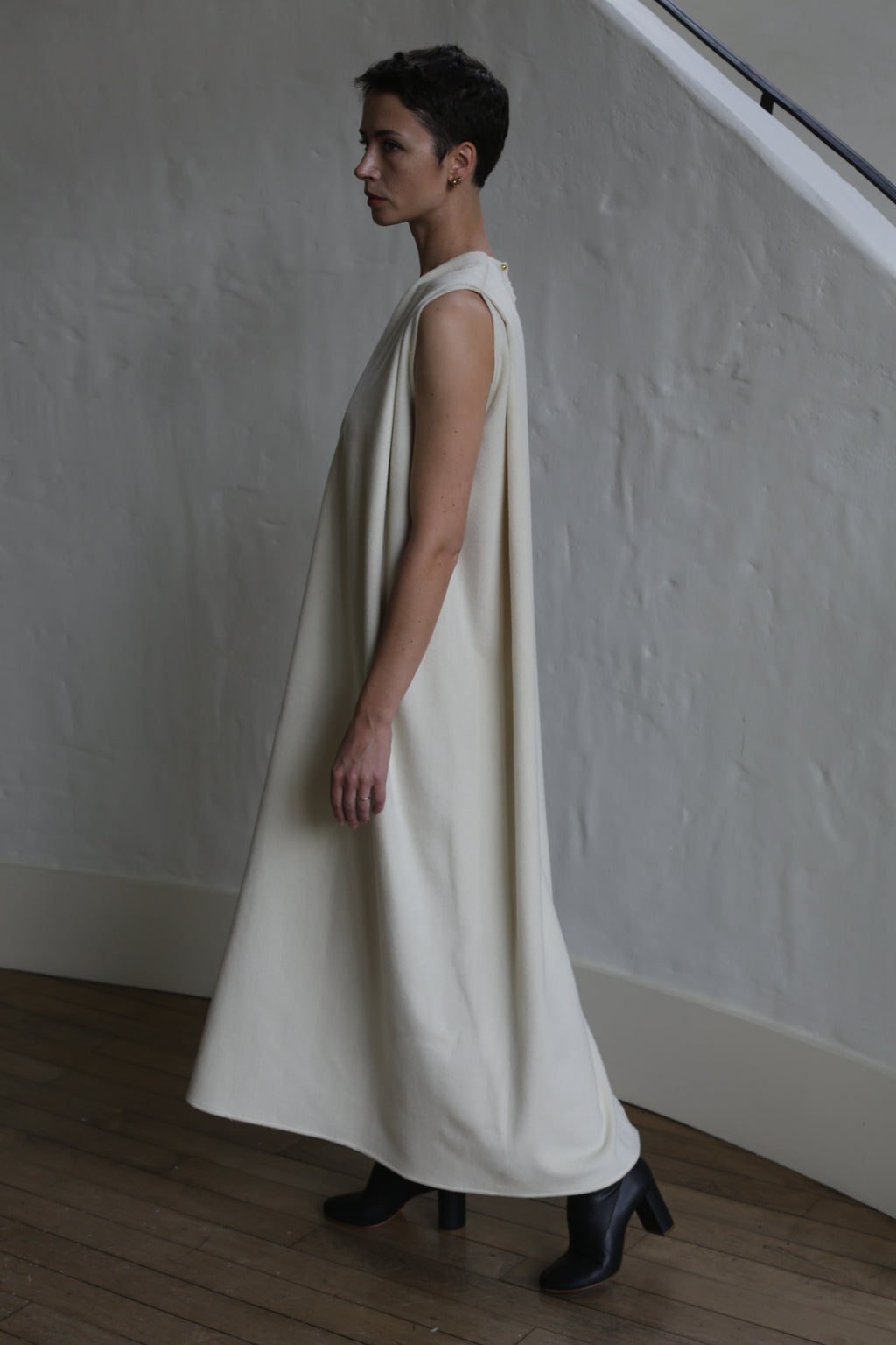 A person with short hair stands in profile, wearing The Winter Dress | Cashmere Undyed— a loose, sleeveless, long cream dress—plus black ankle boots, against a textured white wall and wooden floor.