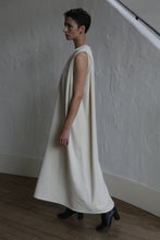Load image into Gallery viewer, A person with short hair stands in profile, wearing The Winter Dress | Cashmere Undyed— a loose, sleeveless, long cream dress—plus black ankle boots, against a textured white wall and wooden floor.