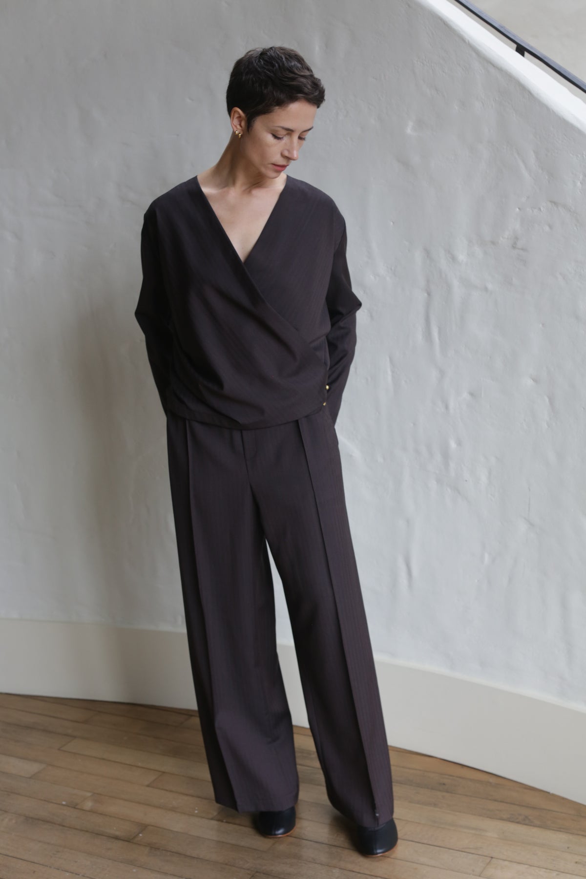 A person with short dark hair stands indoors on wooden flooring, wearing the X Blouse | Wool Herringbone Brown as part of a loose, wide-legged suit, looking down with hands behind their back against a white textured wall.