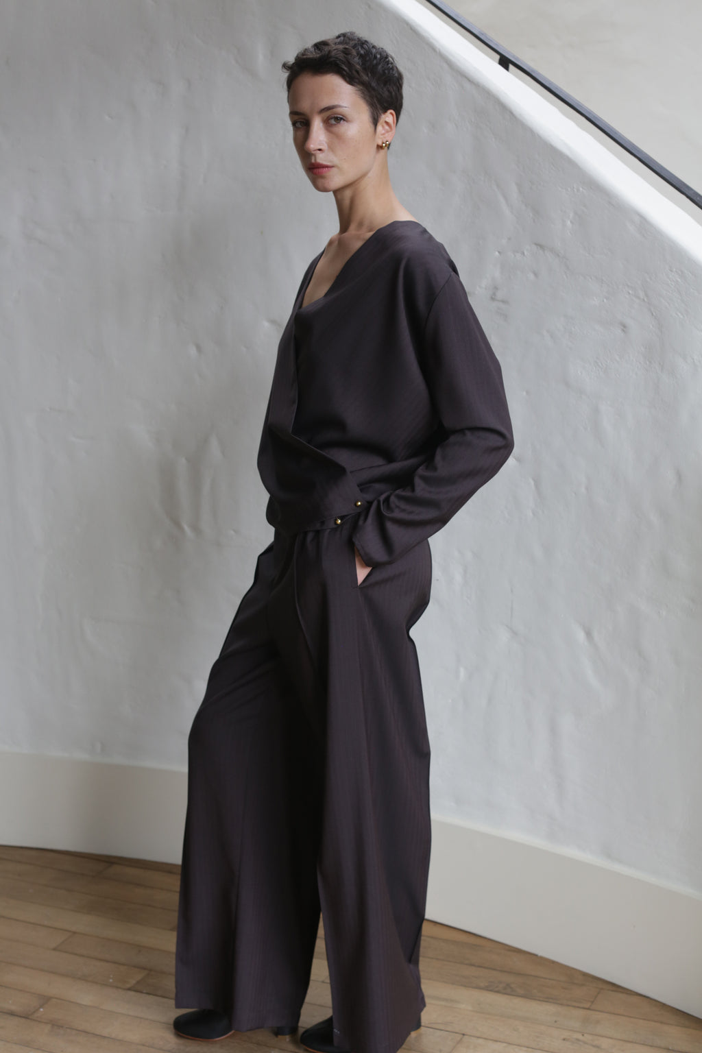 A person with short dark hair stands indoors against a white textured wall, wearing wide-leg Wool Herringbone Trousers in brown and a loose-fitting dark long-sleeve top, one hand in their pocket, looking at the camera.