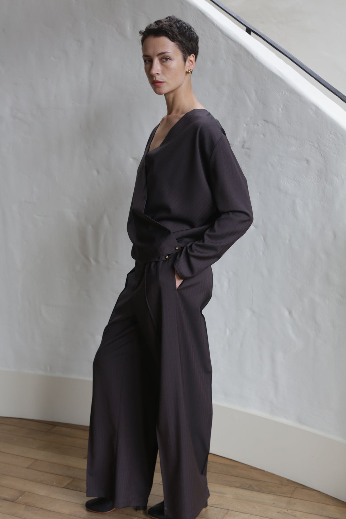 A person with short dark hair stands indoors against a white textured wall, wearing wide-leg Wool Herringbone Trousers in brown and a loose-fitting dark long-sleeve top, one hand in their pocket, looking at the camera.
