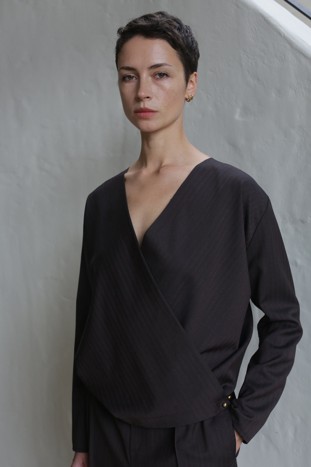 A person with short dark hair, wearing the X Blouse in Wool Herringbone Brown and matching pants, stands against a light textured wall, looking calmly at the camera.