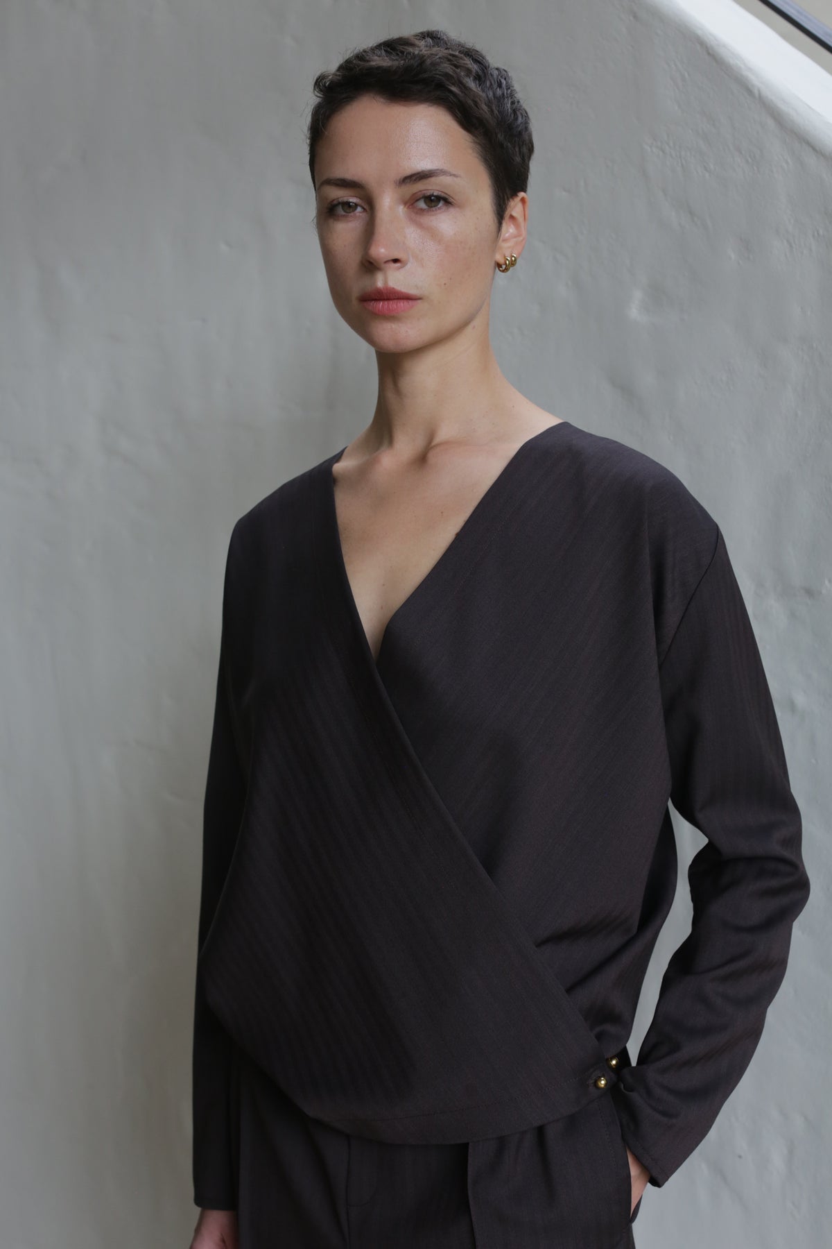 A person with short dark hair, wearing the X Blouse in Wool Herringbone Brown and matching pants, stands against a light textured wall, looking calmly at the camera.