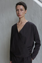 Load image into Gallery viewer, A person with short dark hair, wearing the X Blouse in Wool Herringbone Brown and matching pants, stands against a light textured wall, looking calmly at the camera.