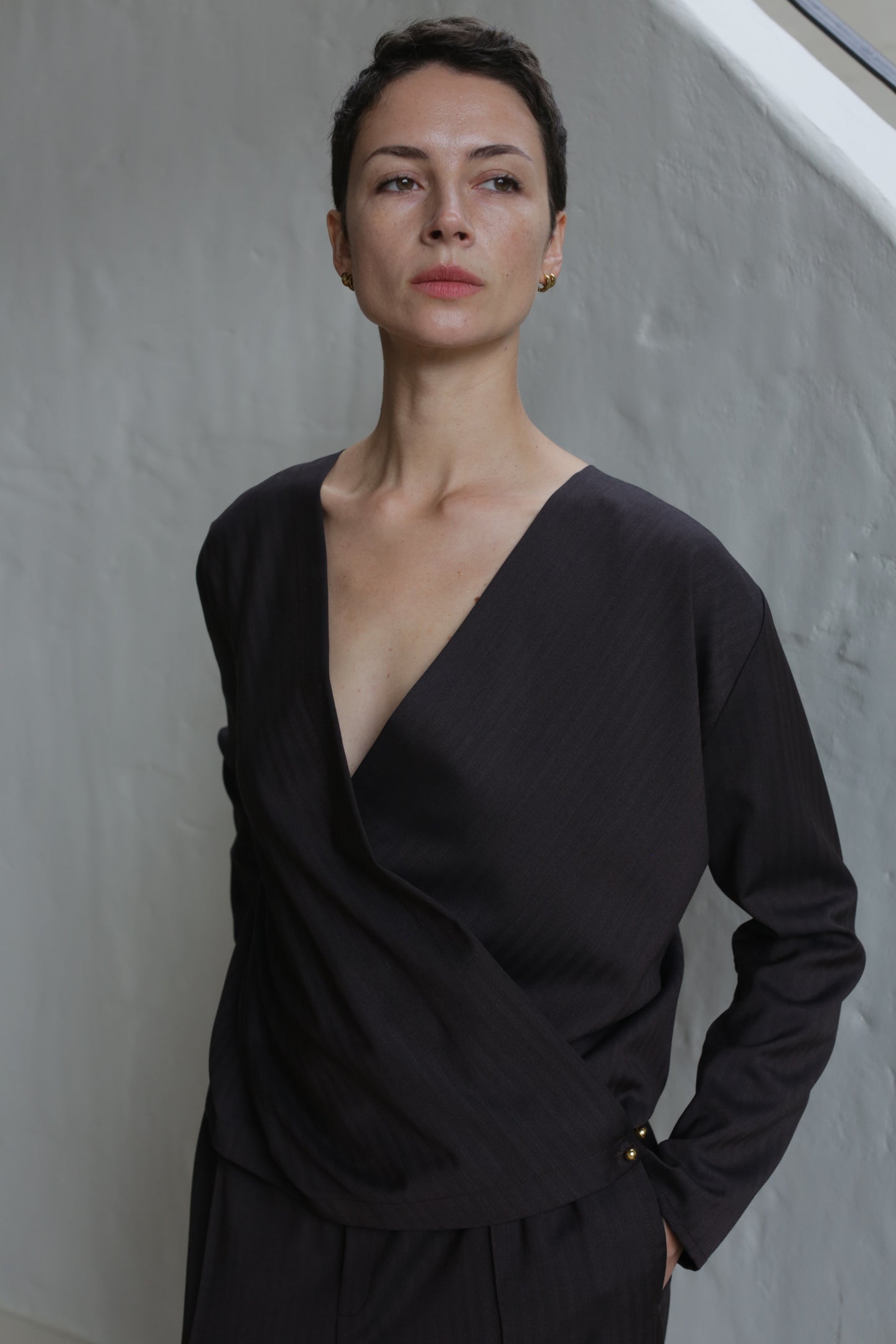 A person with short dark hair, wearing the X Blouse in Wool Herringbone Brown with a deep V-neck, stands against a light-colored wall and gazes slightly upward with a confident expression.
