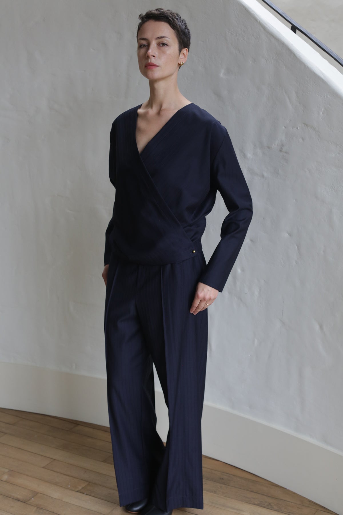 A person with short hair wears the X Blouse in Wool Pinstripe Navy and wide-leg trousers, standing indoors against a white textured wall and wooden floor, looking confidently at the camera.