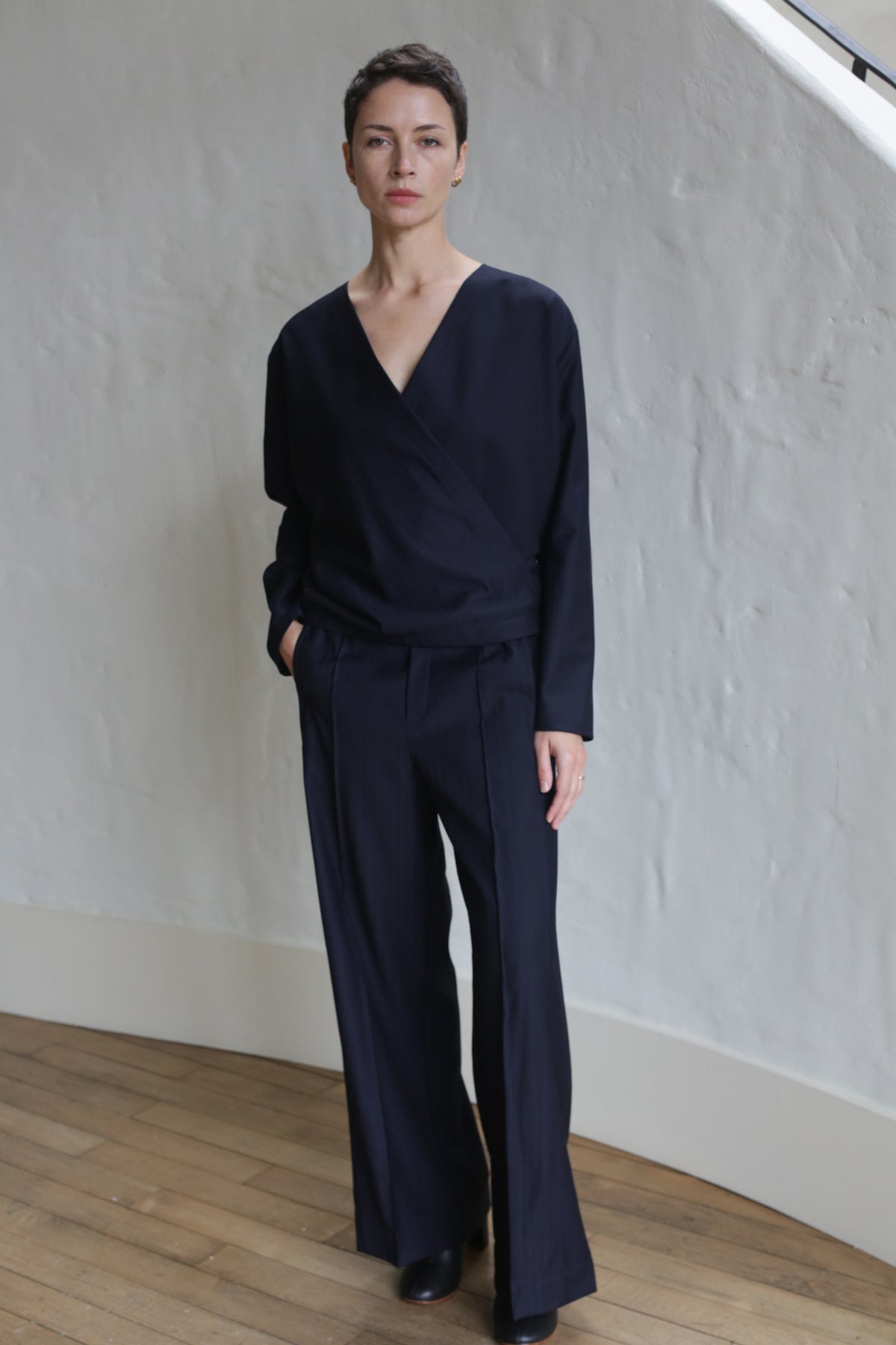 A person with short dark hair stands indoors against a textured white wall, wearing the X Blouse | Wool Pinstripe Navy and matching wide-leg trousers, with one hand in their pocket.