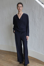 Load image into Gallery viewer, A person with short dark hair stands indoors against a textured white wall, wearing the X Blouse | Wool Pinstripe Navy and matching wide-leg trousers, with one hand in their pocket.