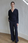 A woman stands indoors against a textured white wall, wearing navy Pinstripe Trousers and a matching loose, long-sleeved wrap top. She has short hair and a neutral expression.