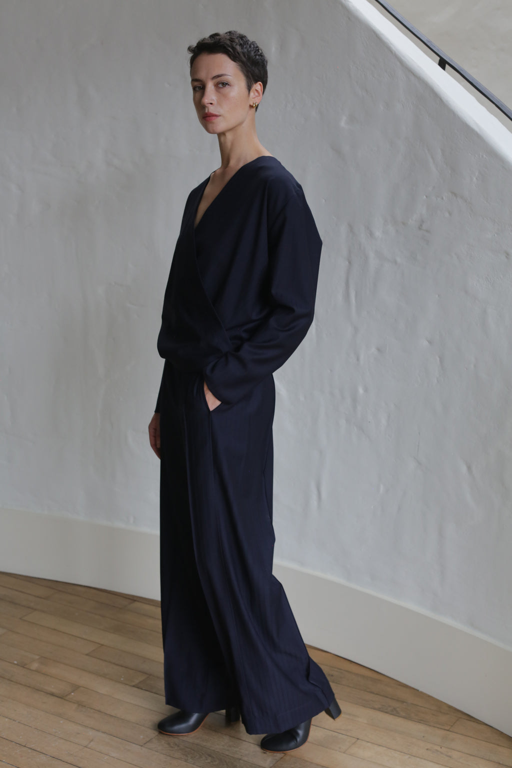 A person with short dark hair wearing navy Pinstripe Trousers, a loose long-sleeve top, and black shoes stands indoors against a textured white wall, looking at the camera with hands in their pockets.