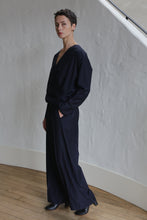 Load image into Gallery viewer, A person with short dark hair wearing navy Pinstripe Trousers, a loose long-sleeve top, and black shoes stands indoors against a textured white wall, looking at the camera with hands in their pockets.