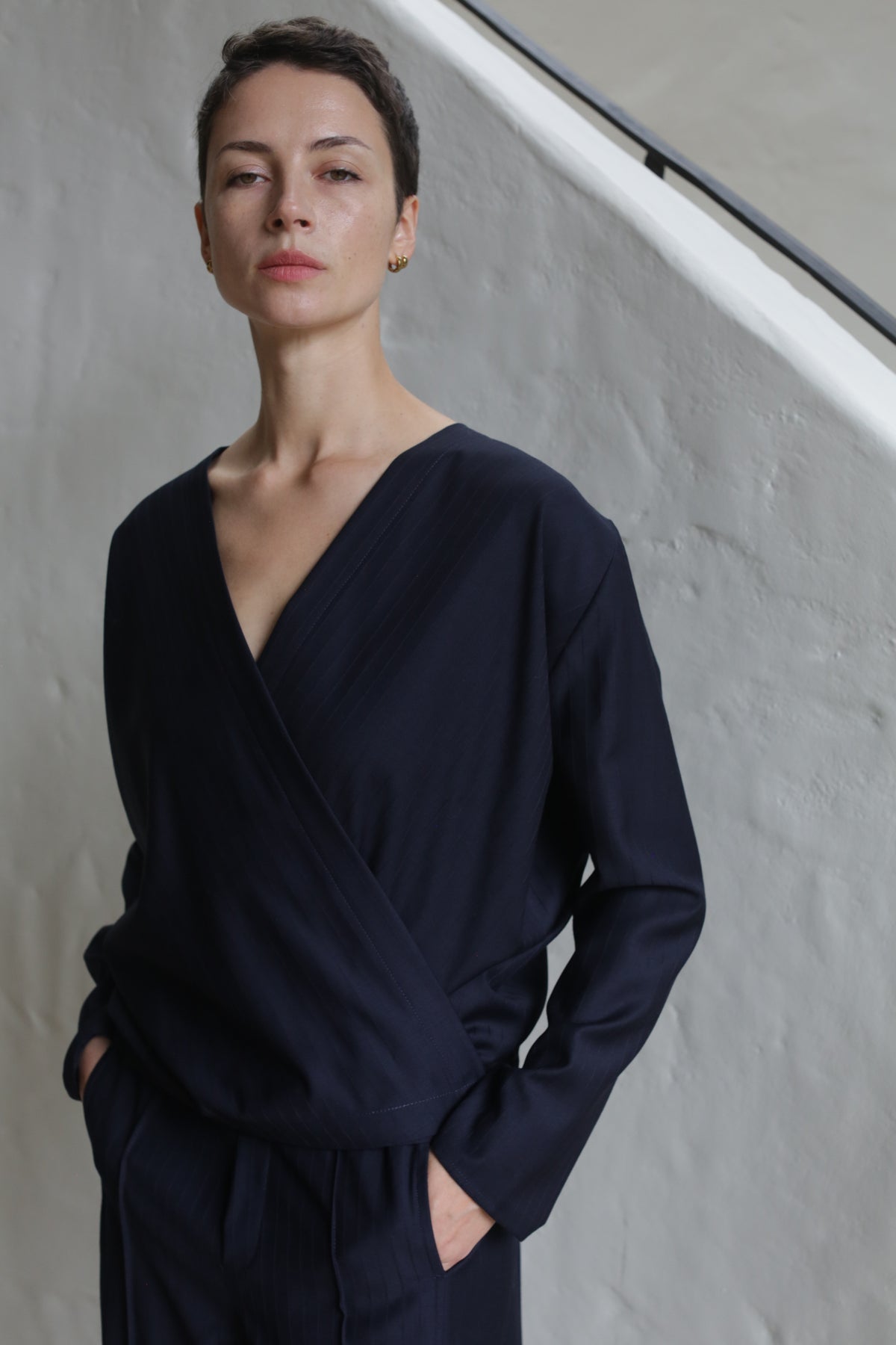 A person with short hair stands with hands in pockets, wearing the X Blouse in Wool Pinstripe Navy and matching pants, against a light textured wall and staircase.