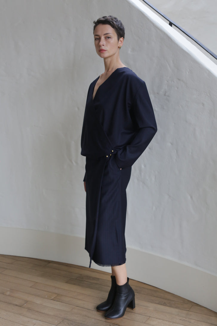 A person with short hair stands indoors near a textured white wall, wearing the Wool Pinstripe Navy Wrap Skirt and black ankle boots, looking confidently at the camera.