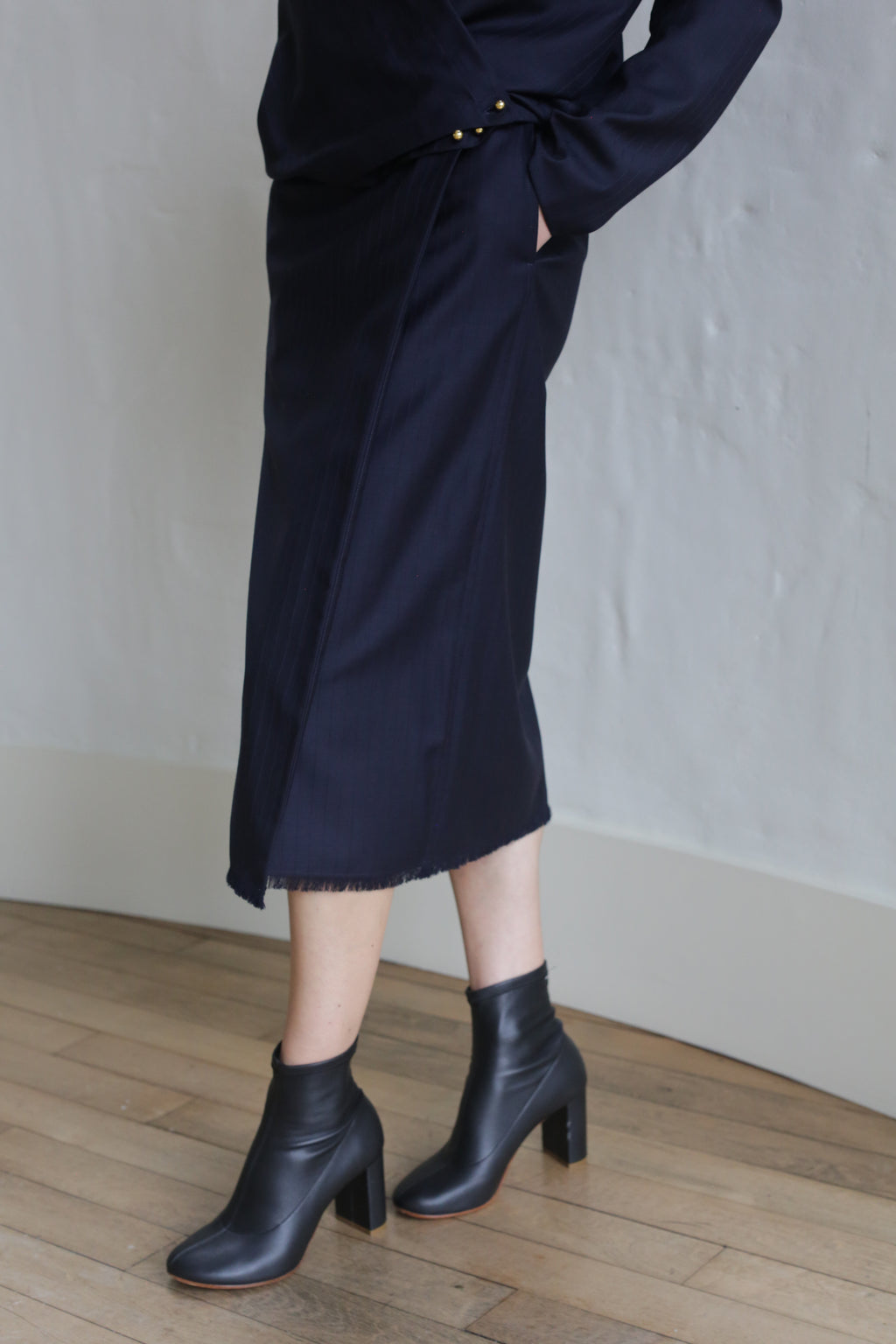 A person wearing the Wrap Skirt | Wool Pinstripe Navy stands on a wooden floor against a light-colored wall, hands in pockets. Black heeled ankle boots complete the look, with the upper body mostly out of frame.