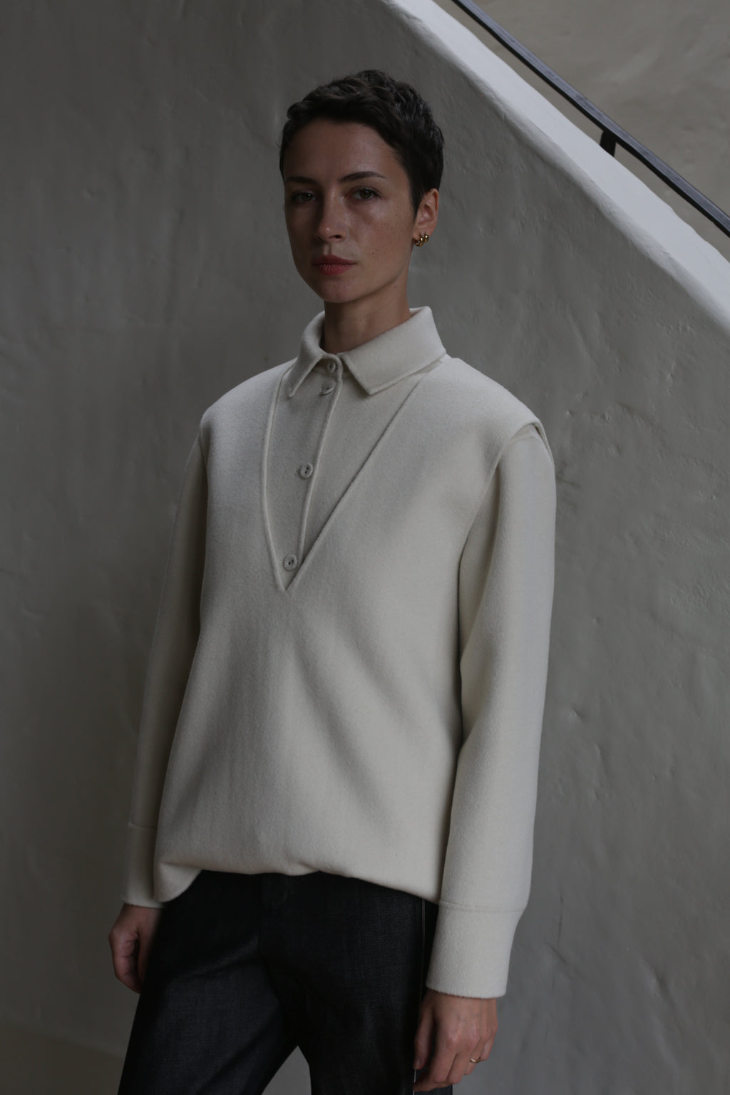 A person with short hair stands indoors against a textured light wall and staircase railing, wearing an undyed cream-colored cashmere vest and dark pants.