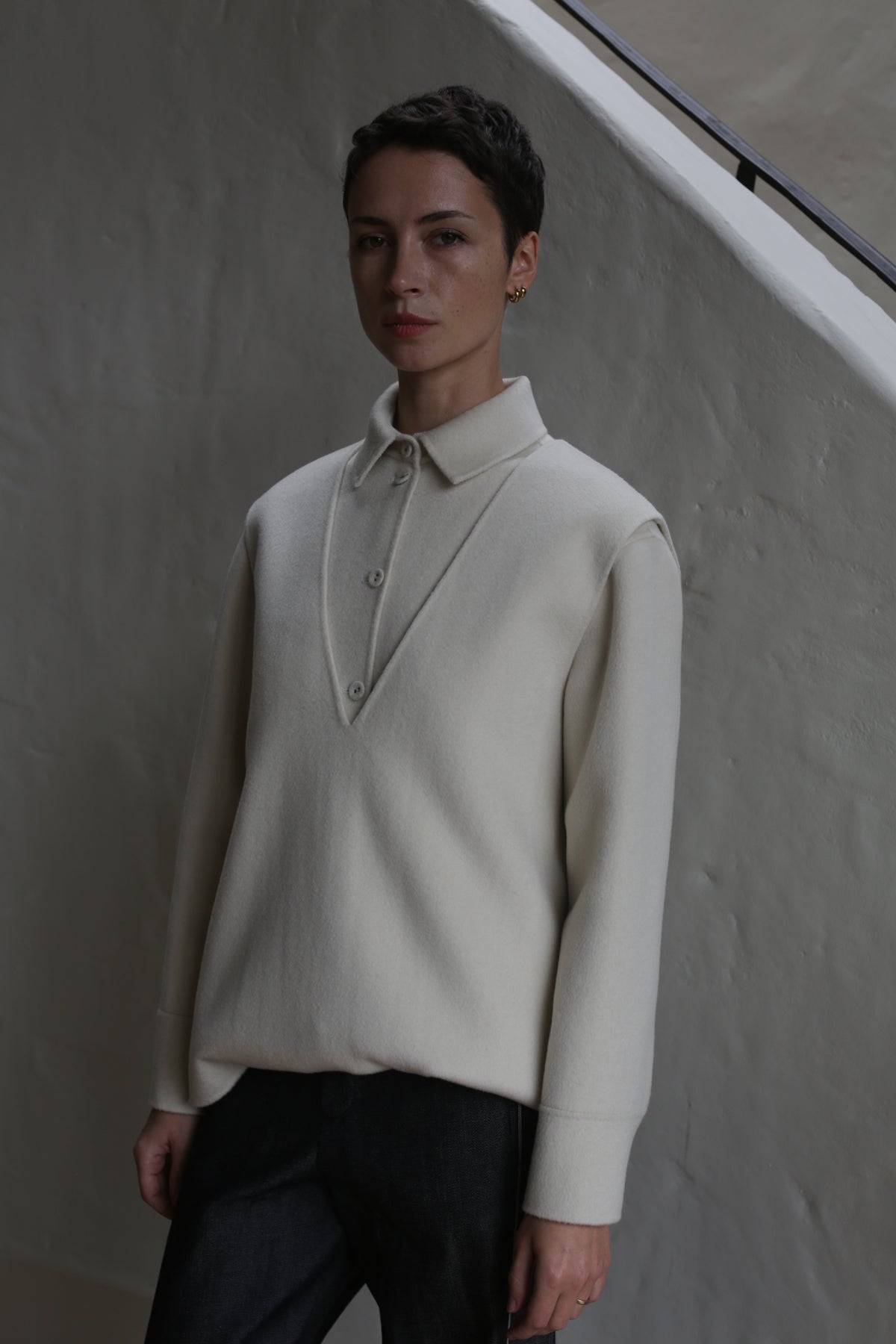 A person with short hair stands indoors against a textured light wall and staircase railing, wearing an undyed cream-colored cashmere vest and dark pants.