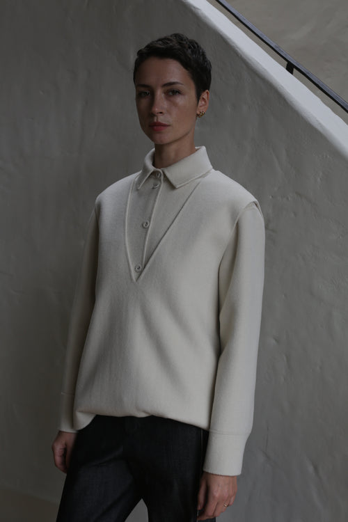 A person with short hair stands indoors against a textured light wall and staircase railing, wearing an undyed cream-colored cashmere vest and dark pants.