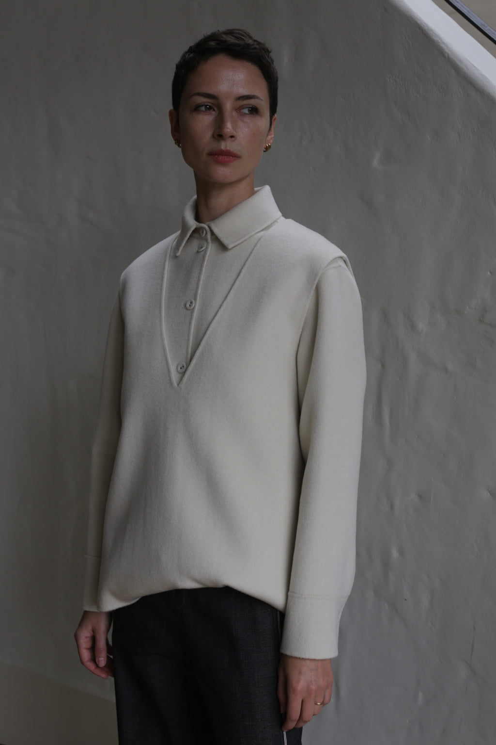 A person with short dark hair wears the undyed Cashmere Vest, paired with dark trousers, standing indoors against a textured wall in soft lighting.