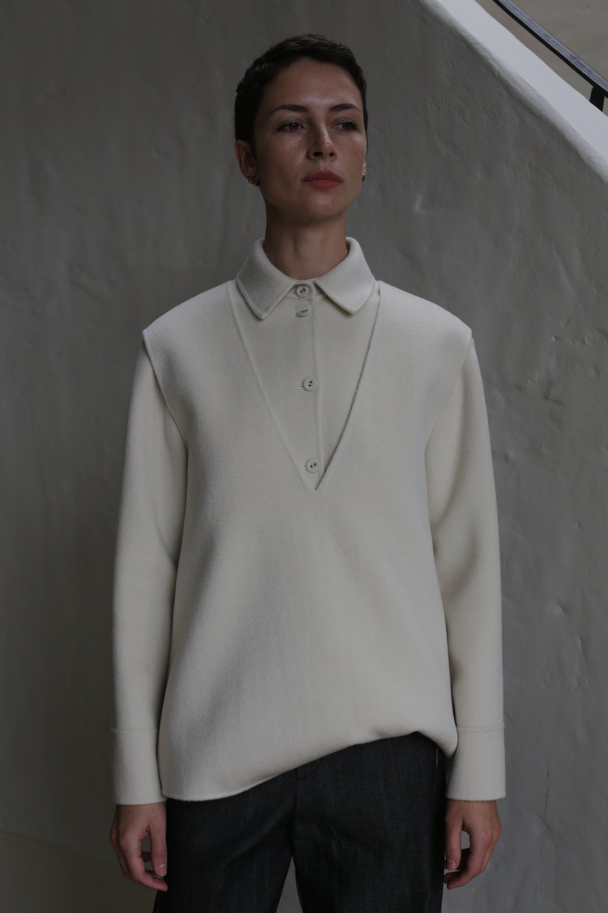 A person with short hair wears a cream-colored, undyed cashmere vest and dark pants, standing against a textured light wall and looking slightly to the side.