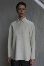 Load image into Gallery viewer, A person with short dark hair wears the Studio Shirt | Undyed, a cream-colored button-up with two front pockets, standing against a textured light gray wall.