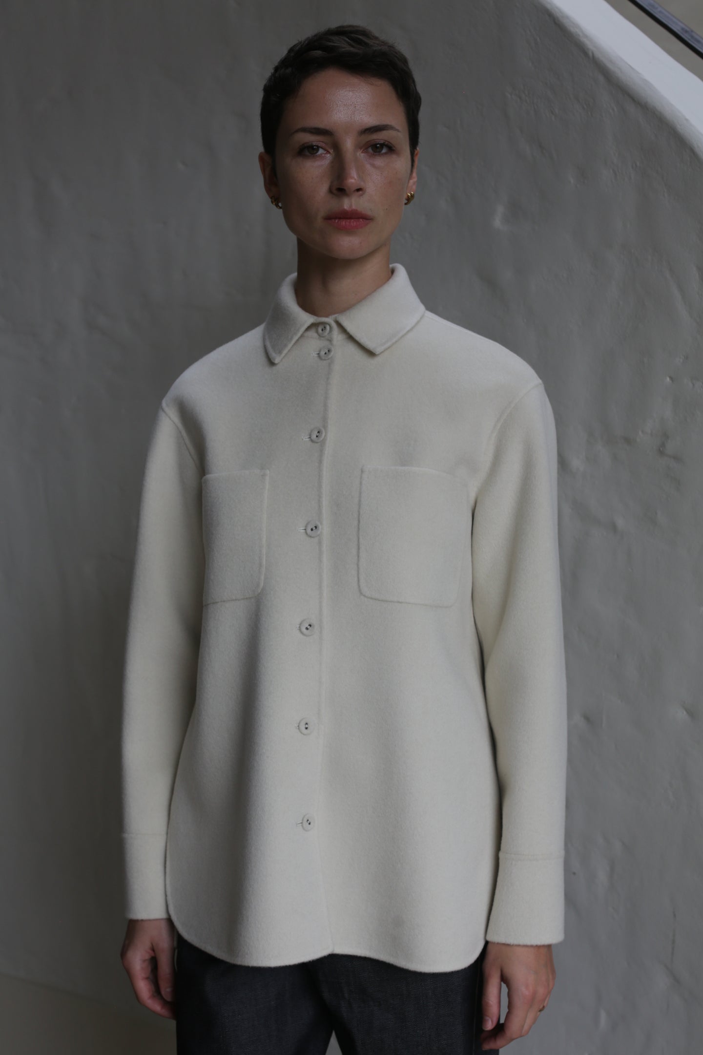 A person with short dark hair wears the Studio Shirt | Undyed, a cream-colored button-up with two front pockets, standing against a textured light gray wall.