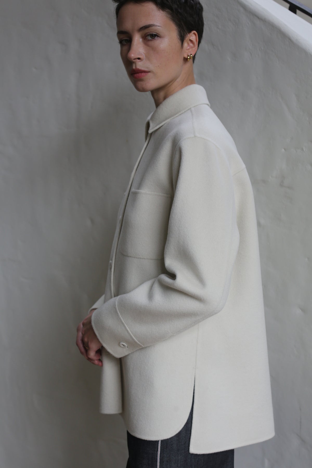 A person with short dark hair wears the Studio Shirt | Undyed and dark pants, standing sideways against a light background, looking at the camera with a neutral expression.