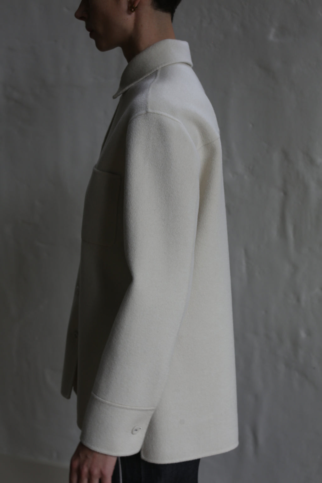 A person stands in profile against a textured gray wall, wearing the Studio Shirt | Undyed. Soft lighting casts subtle shadows, highlighting the smooth fabric and classic long-sleeved design of this light-colored shirt.