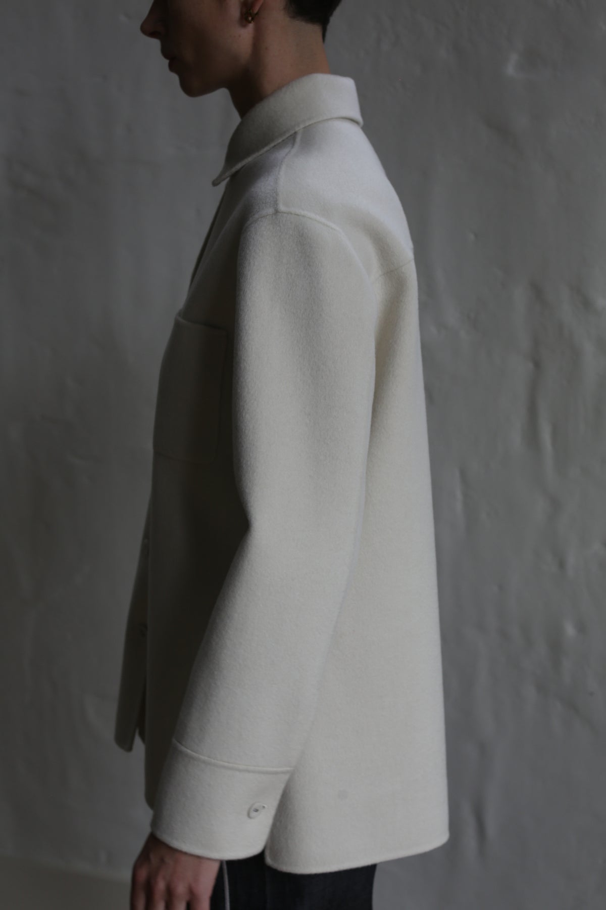 A person stands in profile against a textured gray wall, wearing the Studio Shirt | Undyed. Soft lighting casts subtle shadows, highlighting the smooth fabric and classic long-sleeved design of this light-colored shirt.