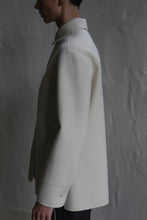 Load image into Gallery viewer, A person stands in profile against a textured gray wall, wearing the Studio Shirt | Undyed. Soft lighting casts subtle shadows, highlighting the smooth fabric and classic long-sleeved design of this light-colored shirt.