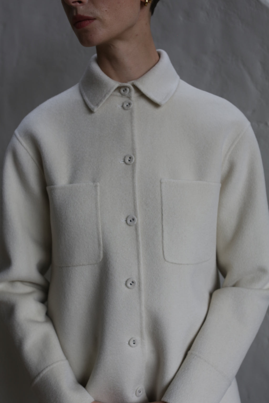 A person models the Studio Shirt | Undyed, a cream-colored wool button-up with two front pockets, standing against a gray textured background. Their hands are loosely clasped, and their face is partially out of frame.