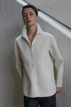 Load image into Gallery viewer, A person with short hair models the Studio Shirt | Undyed, a white button-up with front pockets, standing against a light-colored wall and looking confidently at the camera.