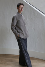 Load image into Gallery viewer, A person with short dark hair stands indoors near a textured white wall, wearing the Studio Shirt in Undyed Natural with their hands in their pockets and paired with dark wide-leg jeans featuring a white stripe on the sides.