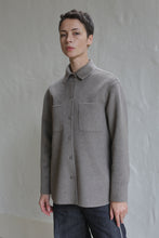 Load image into Gallery viewer, A person with short dark hair wears the Studio Shirt in Undyed Natural, featuring two chest pockets, standing calmly before a light-colored textured wall and looking at the camera.