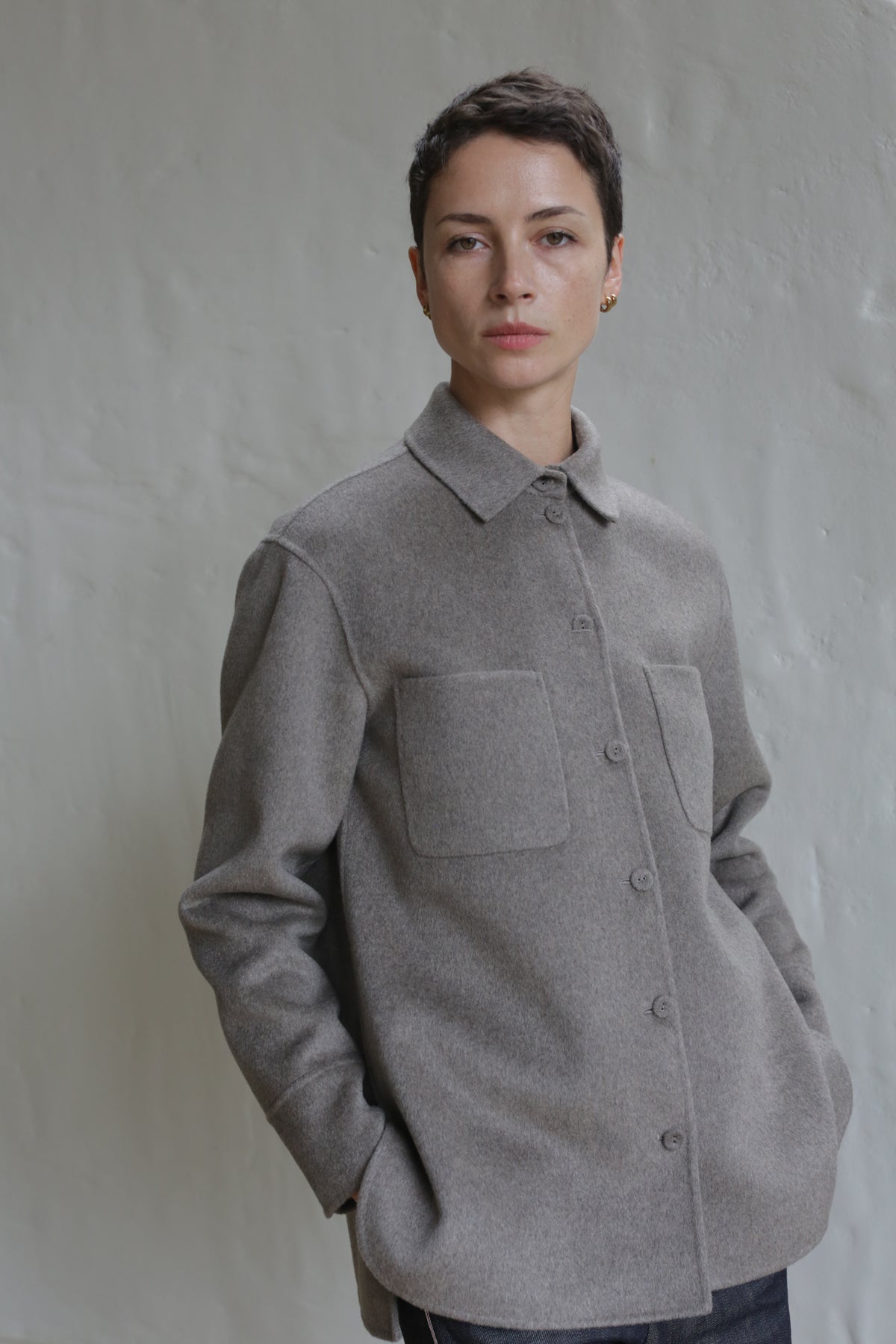 Person with short dark hair wears the Unisex Studio Shirt in Double Face Cashmere Undyed Natural—a luxury button-up with large front pockets—standing against a light gray textured wall, hands in their pockets.