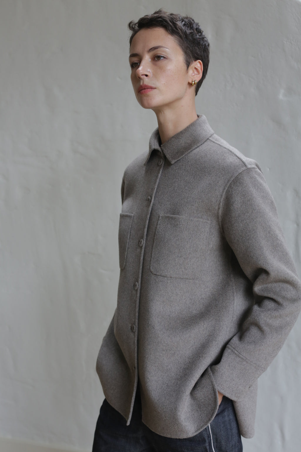 A person with short dark hair wears the Unisex Studio Shirt in Double Face Cashmere Undyed Natural, featuring front pockets, standing against a textured light wall with hands in their pockets, gazing slightly to the side.