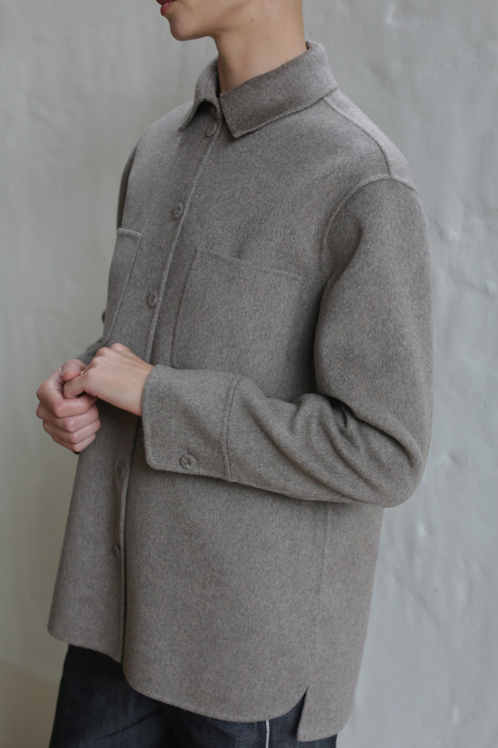A person wearing the Unisex Studio Shirt in Double Face Cashmere Undyed Natural, featuring long sleeves and a collar, stands against a light gray textured wall. Only their upper body is visible; their face is partially out of frame.