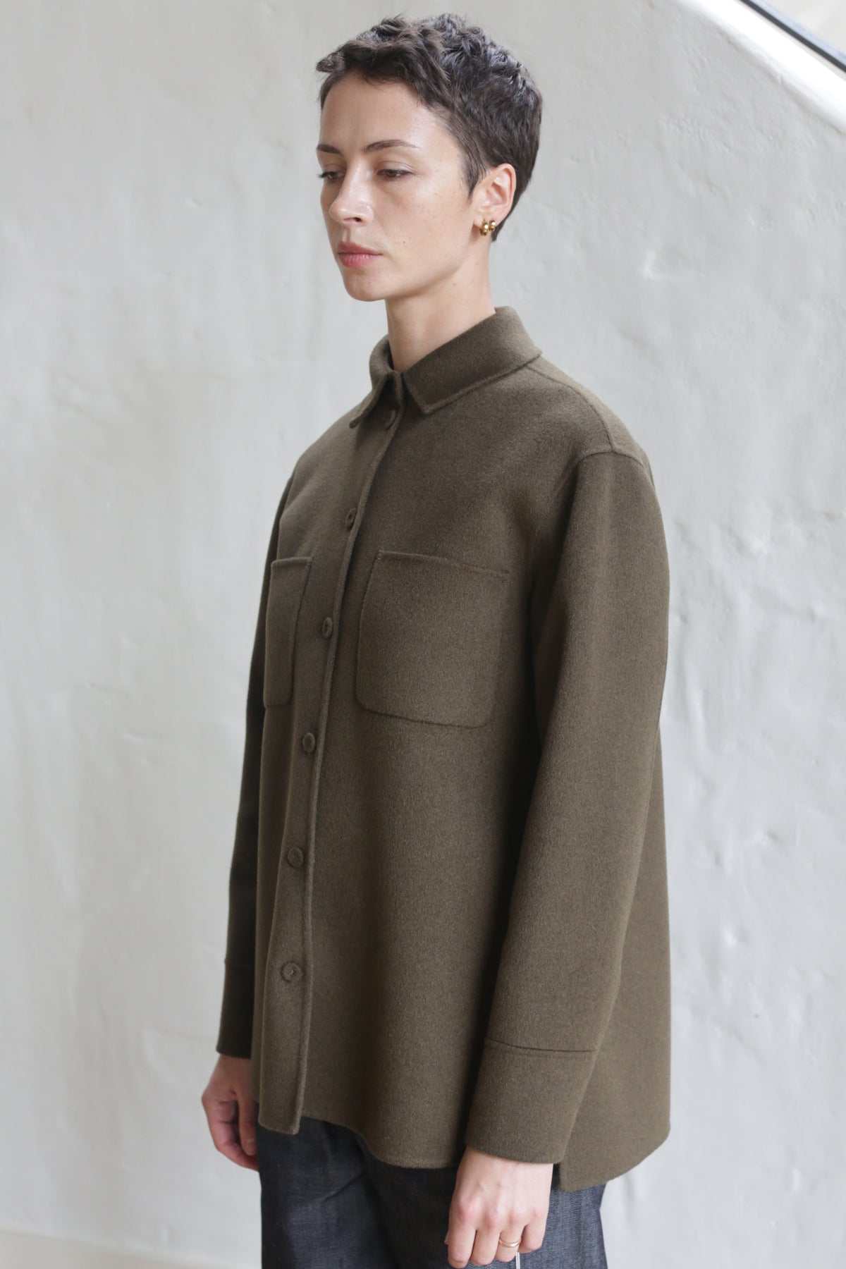 A person with short hair wears the Studio Shirt | Olive, featuring chest pockets, and stands against a white textured wall, looking slightly down and to the side.