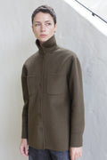 A person with short hair wearing the Studio Shirt in olive, featuring a high collar, buttoned front, and two chest pockets, stands indoors against a white textured wall.
