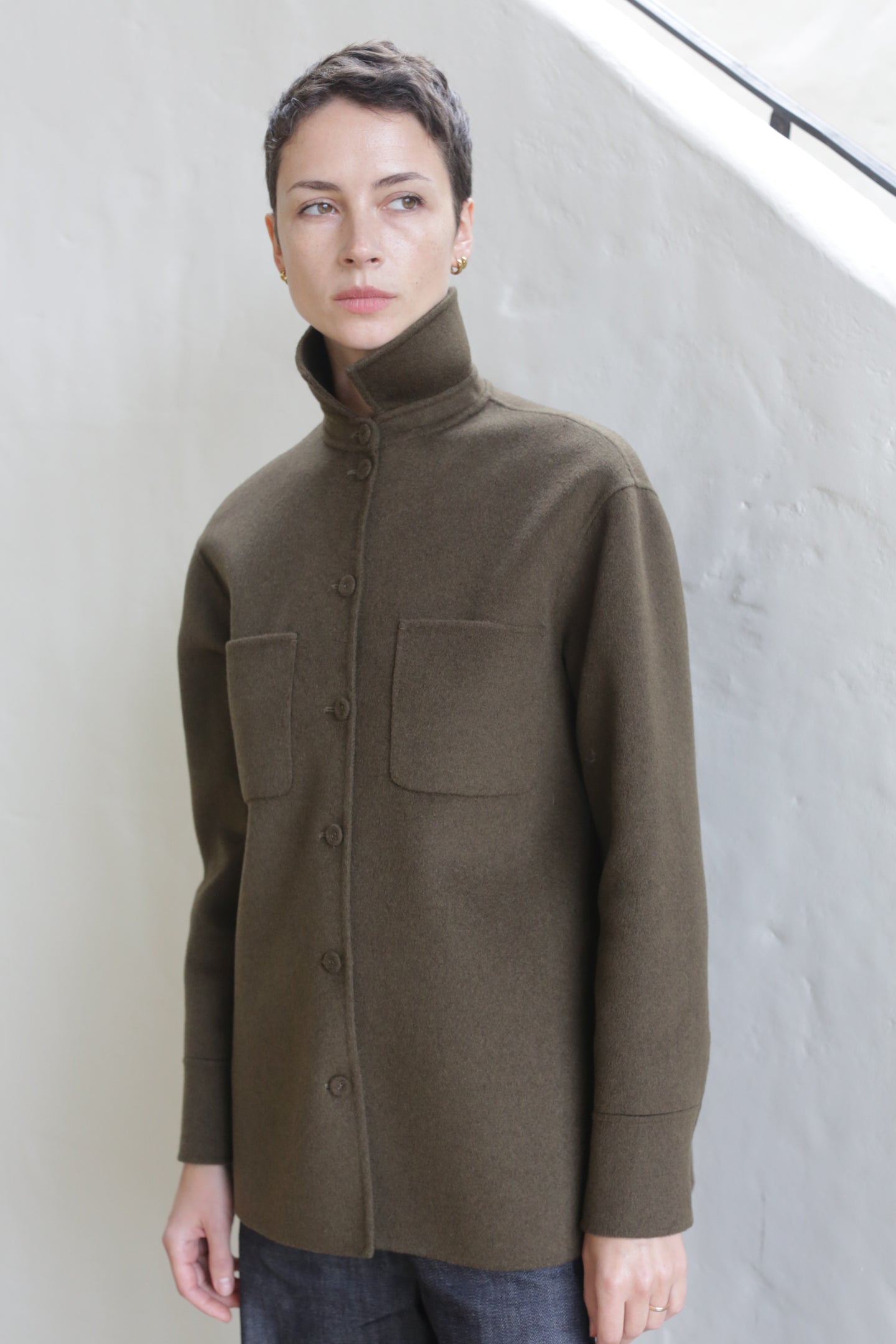 A person with short hair wearing the Studio Shirt in olive, featuring a high collar, buttoned front, and two chest pockets, stands indoors against a white textured wall.