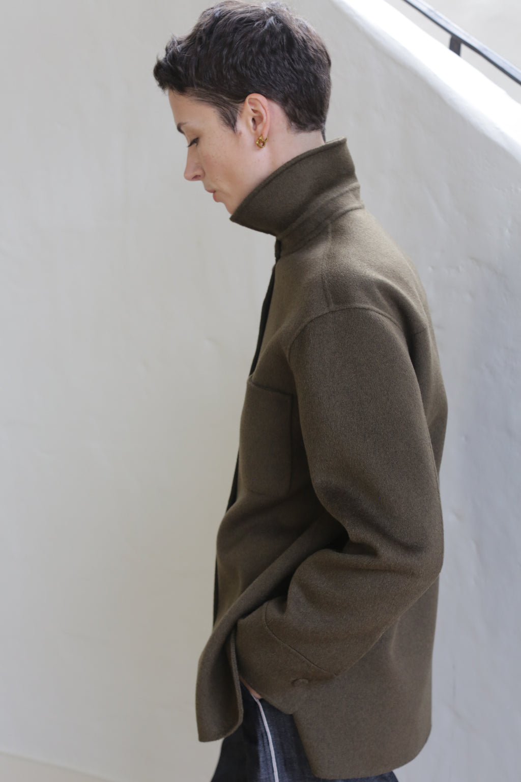 A person with short dark hair stands in profile against a light background, looking down with one hand in their pocket, wearing the Studio Shirt in Olive.