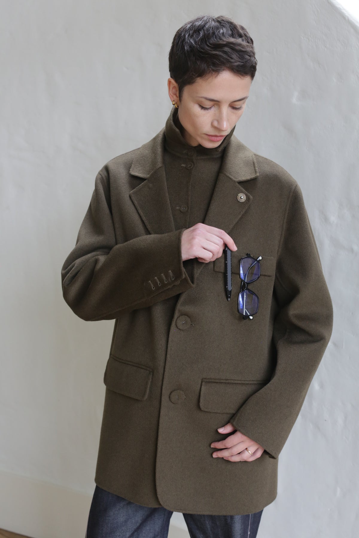 A person with short hair wears the Sculptor's Jacket in Olive over a matching turtleneck, standing against a light wall and looking down while holding eyeglasses and a pen in their breast pocket.