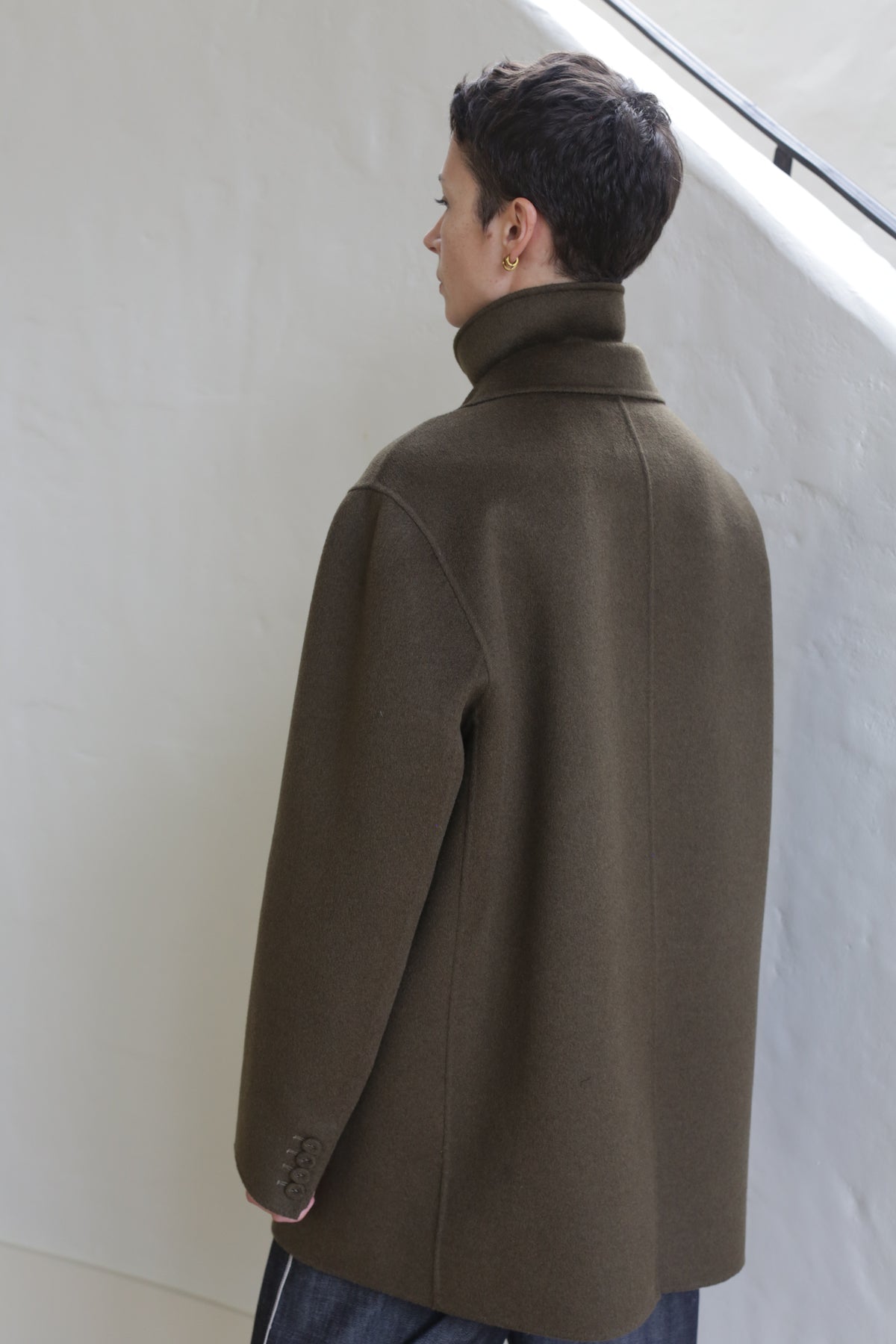 A person with short dark hair, wearing the Sculptor's Jacket in Olive, stands indoors facing away from the camera near a white textured wall and staircase railing.