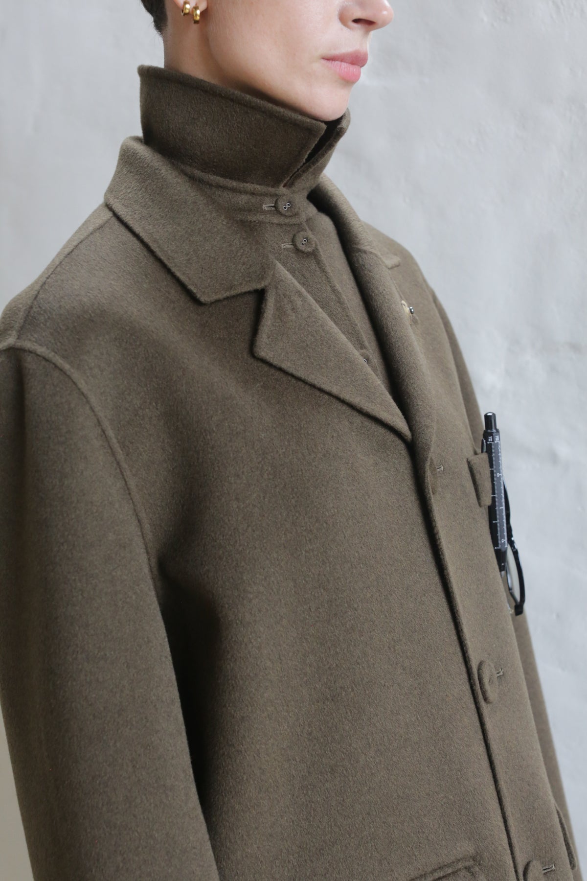 A person wears the Sculptor's Jacket | Olive, a high-collared wool coat, buttoned up with gold earrings and a black accessory clipped to the chest pocket, against a plain light background.