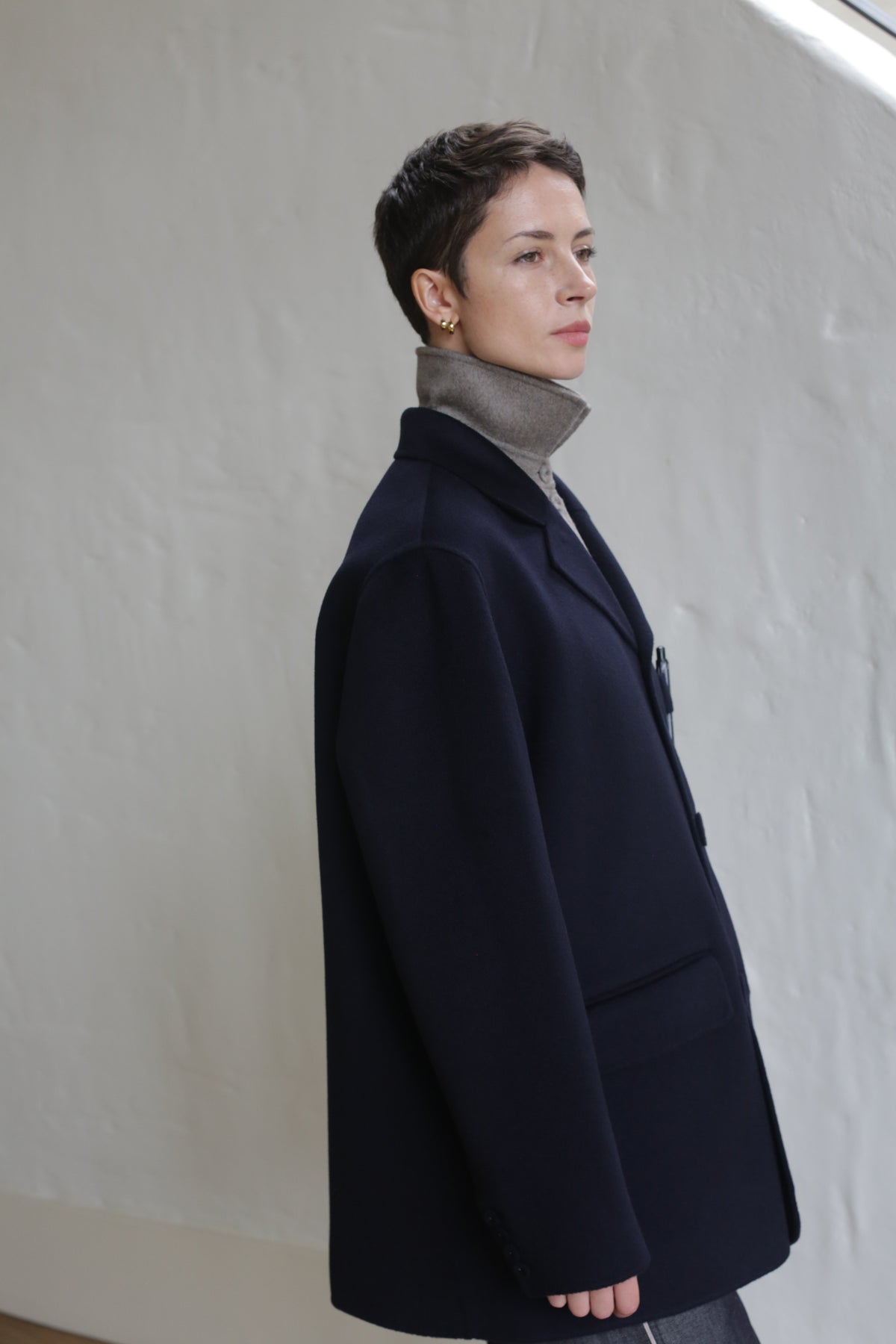 A person with short hair wears the Sculptor's Jacket in Double Face Cashmere Navy over a taupe turtleneck, standing indoors against a light-colored textured wall, gazing thoughtfully into the distance.