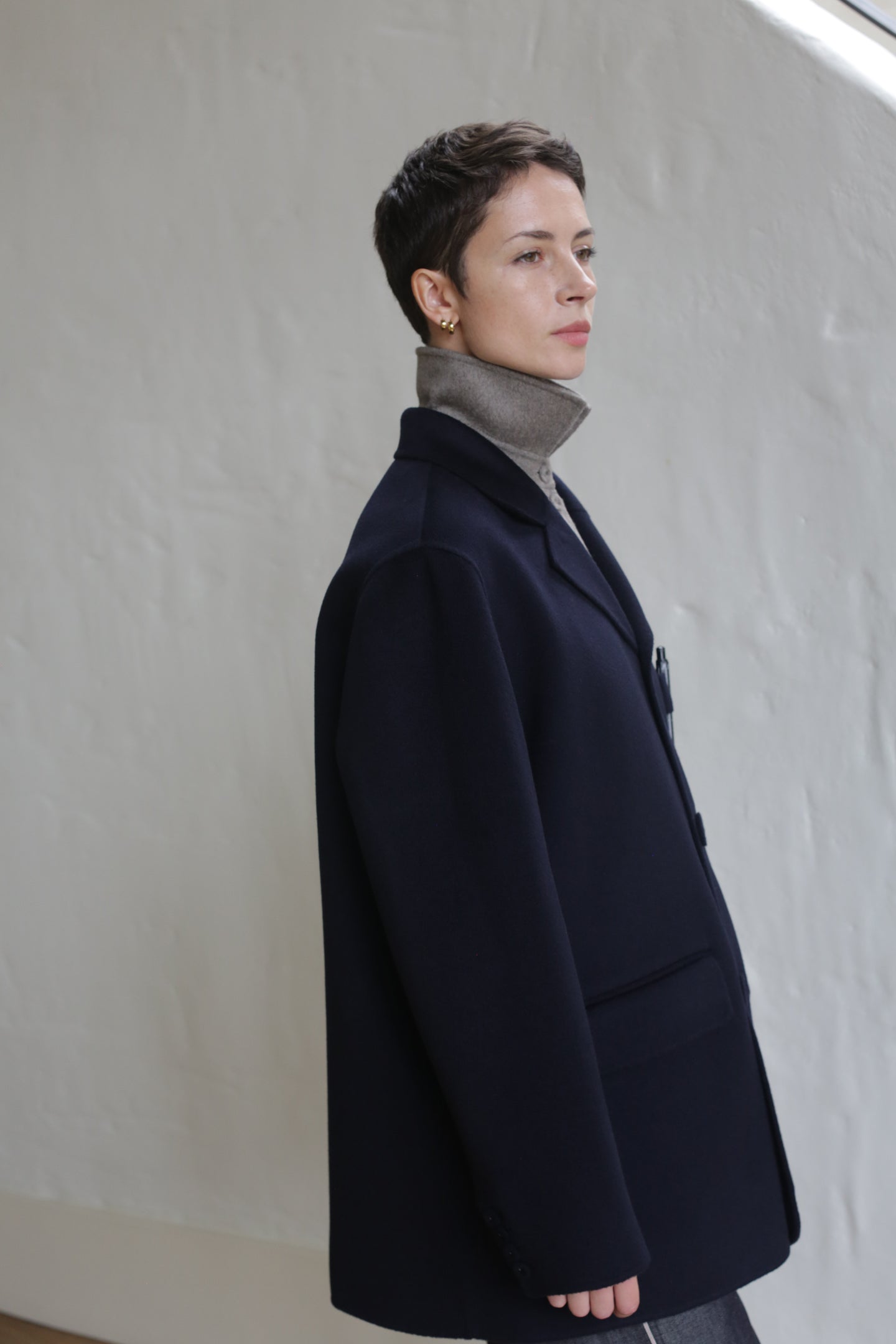 A person with short hair stands in profile, thoughtfully gazing ahead while wearing the Sculptor's Jacket | Navy over a light turtleneck. The plain white wall behind them is softly lit by natural light from above.