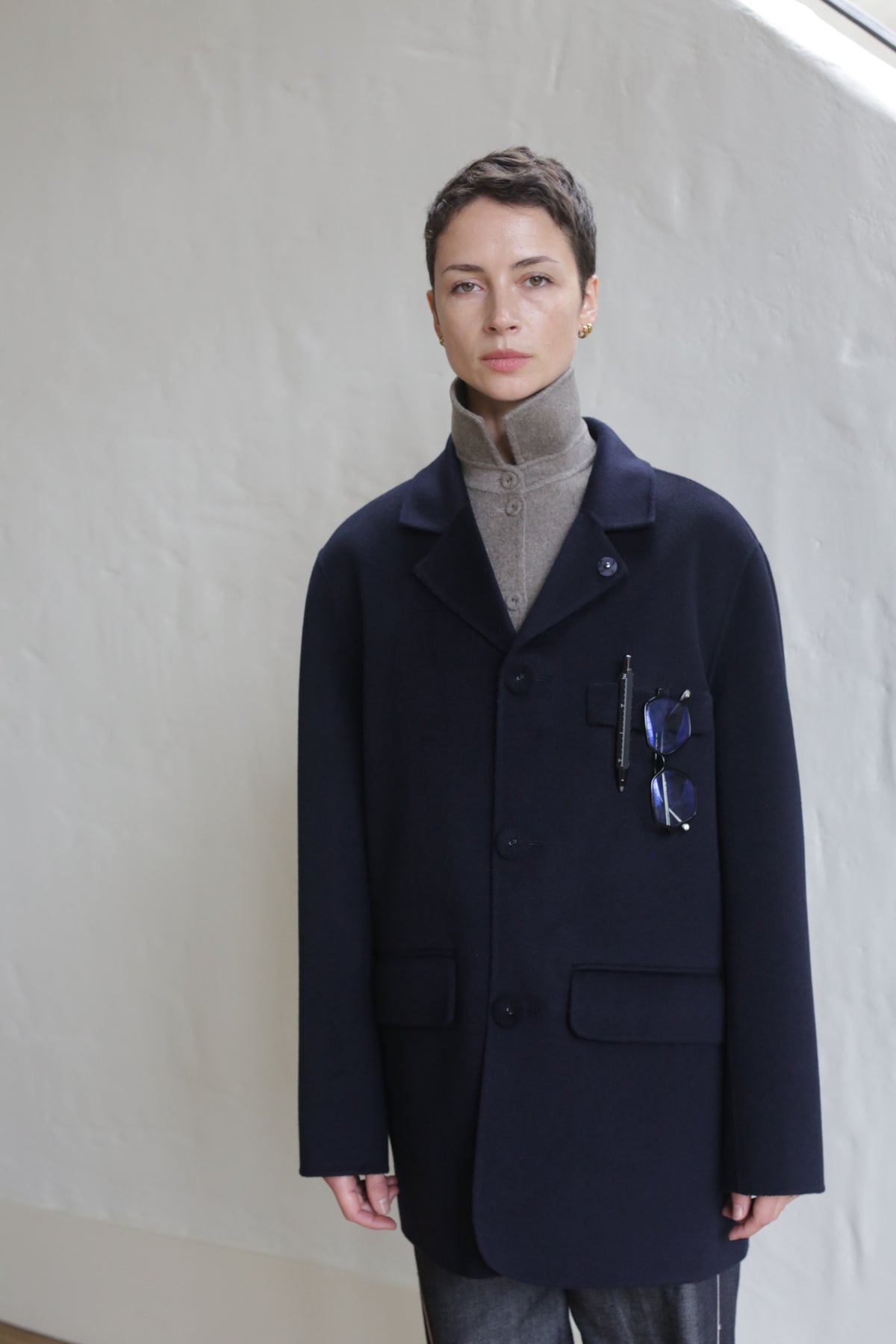 A person with short hair wears the Sculptor's Jacket in Double Face Cashmere Navy over a taupe turtleneck, standing indoors near a light wall. Sunglasses and a pen are clipped to the practical pocket as they look calmly at the camera.