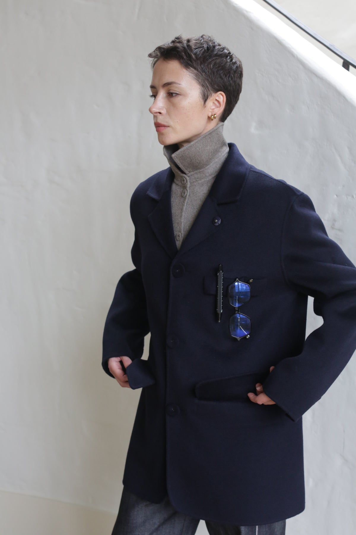 A person with short hair wears the Sculptor's Jacket in double face cashmere navy over a beige turtleneck, blue sunglasses clipped to the pocket. They stand indoors near a white wall, gazing sideways with a composed expression.