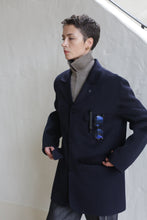Load image into Gallery viewer, A person with short hair wears the Sculptor&#39;s Jacket in navy over a taupe turtleneck, with blue sunglasses clipped to the pocket. They stand against a light-colored wall, gazing off to the side.