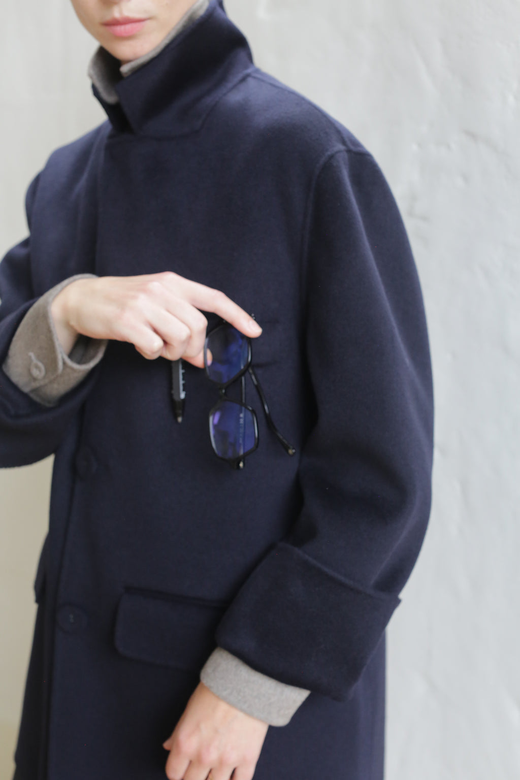 A person in the Sculptor's Jacket | Double Face Cashmere Navy with light gray sleeves holds black-rimmed eyeglasses against a textured background, their face partially out of the frame.